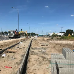 Budowa drogi z kostki brukowej: widok na krawężnik, stos kostki brukowej, ciężarówkę i koparkę Komatsu w tle, na tle błękitnego nieba.