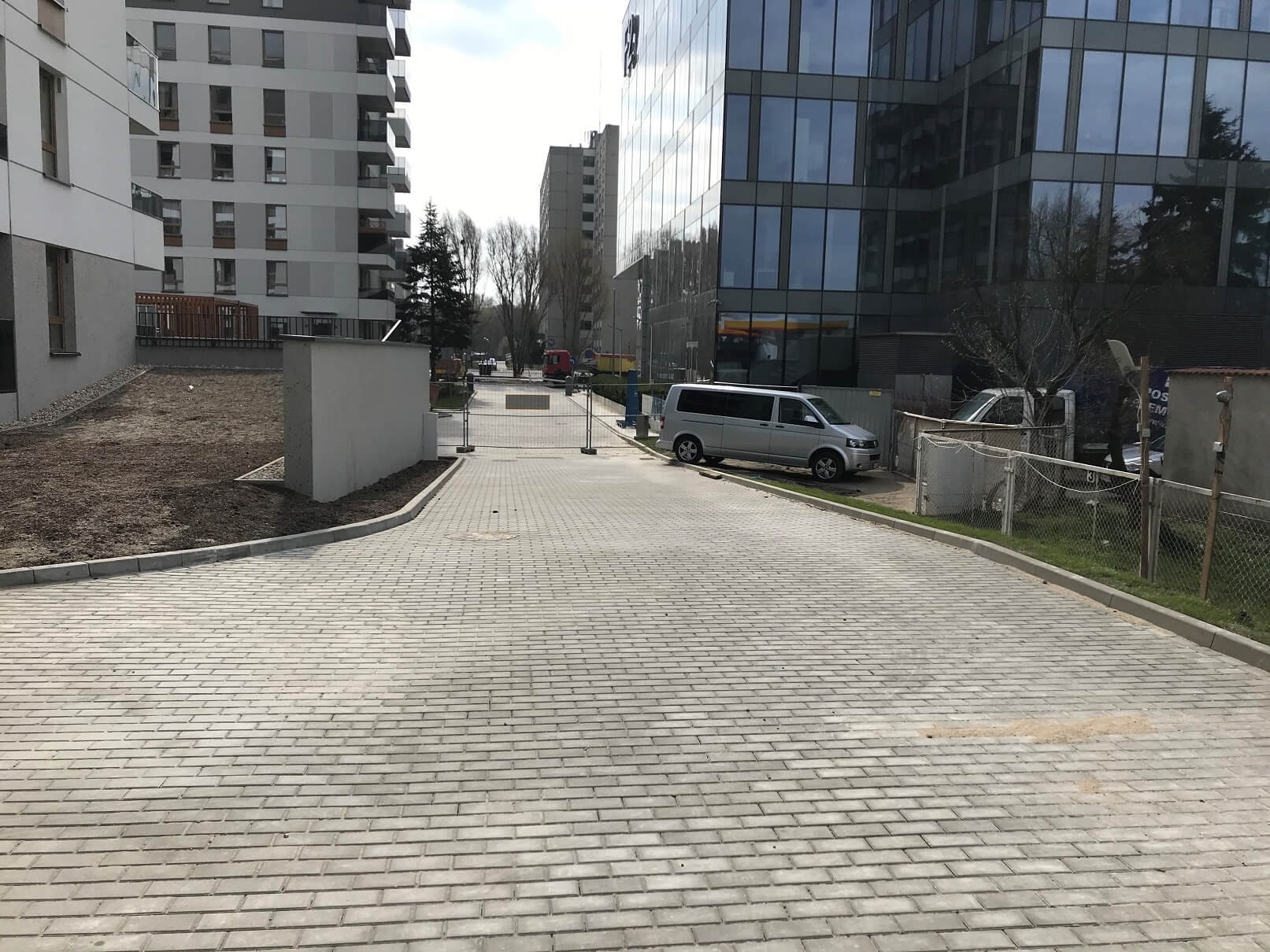 Nowa, szara nawierzchnia z kostki brukowej prowadząca do nowoczesnych budynków biurowych i mieszkalnych, z zaparkowanym srebrnym vanem po prawej stronie.