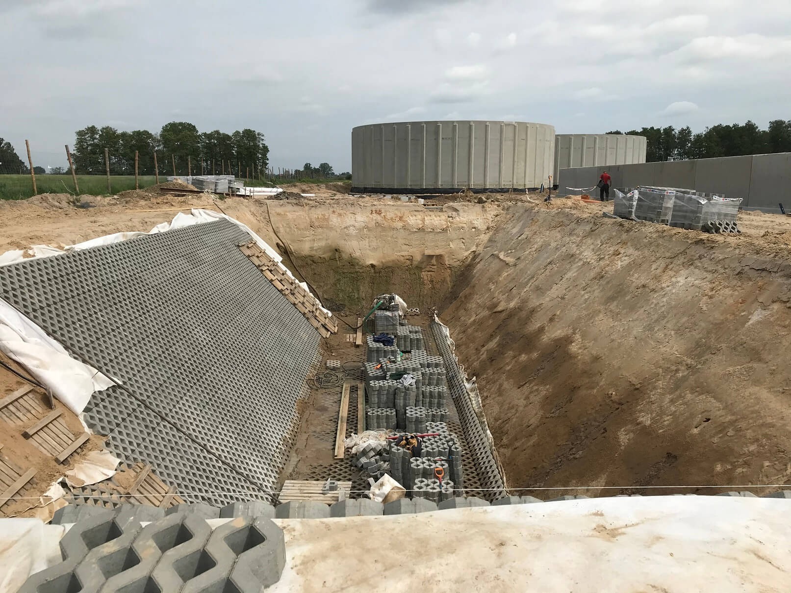 Wykop z zabezpieczoną skarpą z ażurowych płyt betonowych, przygotowany pod budowę zbiorników betonowych widocznych w tle, na placu budowy z ziemistym podłożem.