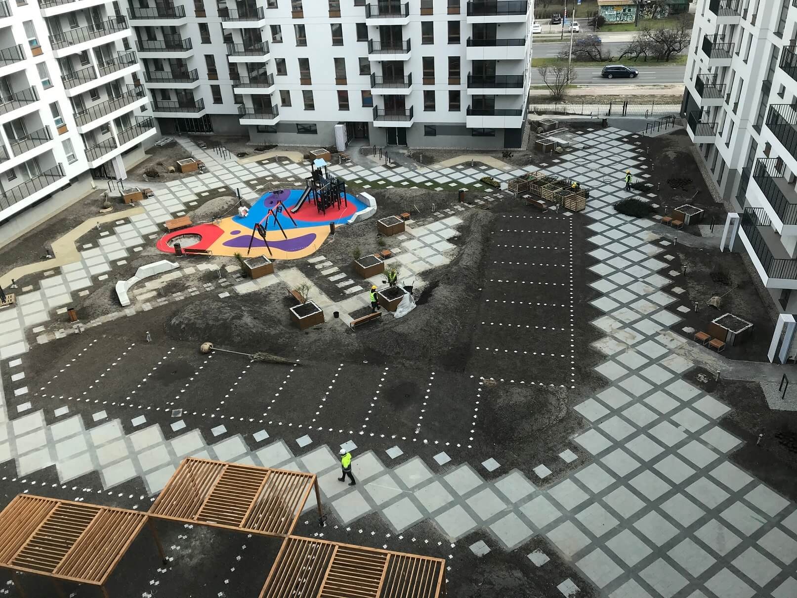Widok z góry na plac budowy osiedla mieszkaniowego z elementami placu zabaw i nową nawierzchnią z kostki w geometryczny wzór, pracownicy w żółtych kamizelkach.