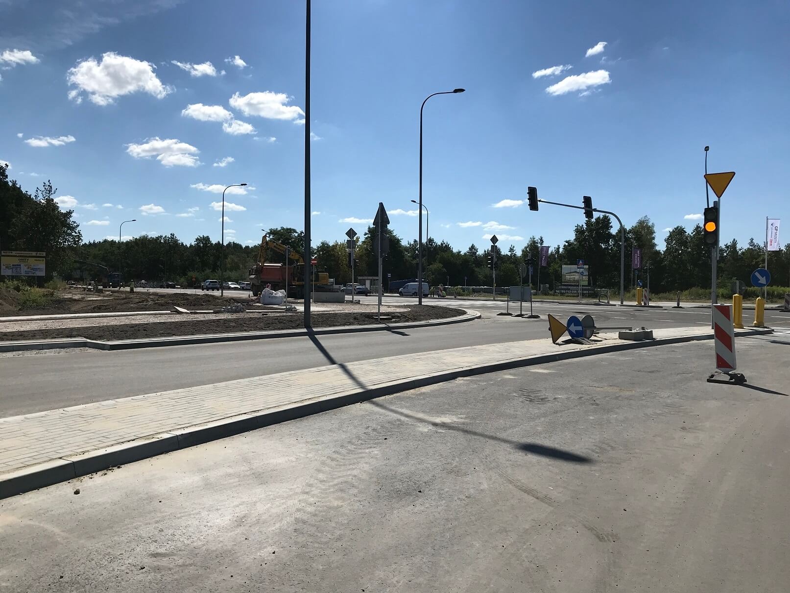 Nowo wybudowane rondo z chodnikiem z kostki brukowej, sygnalizacją świetlną i znakami drogowymi, w tle widoczny sprzęt budowlany.