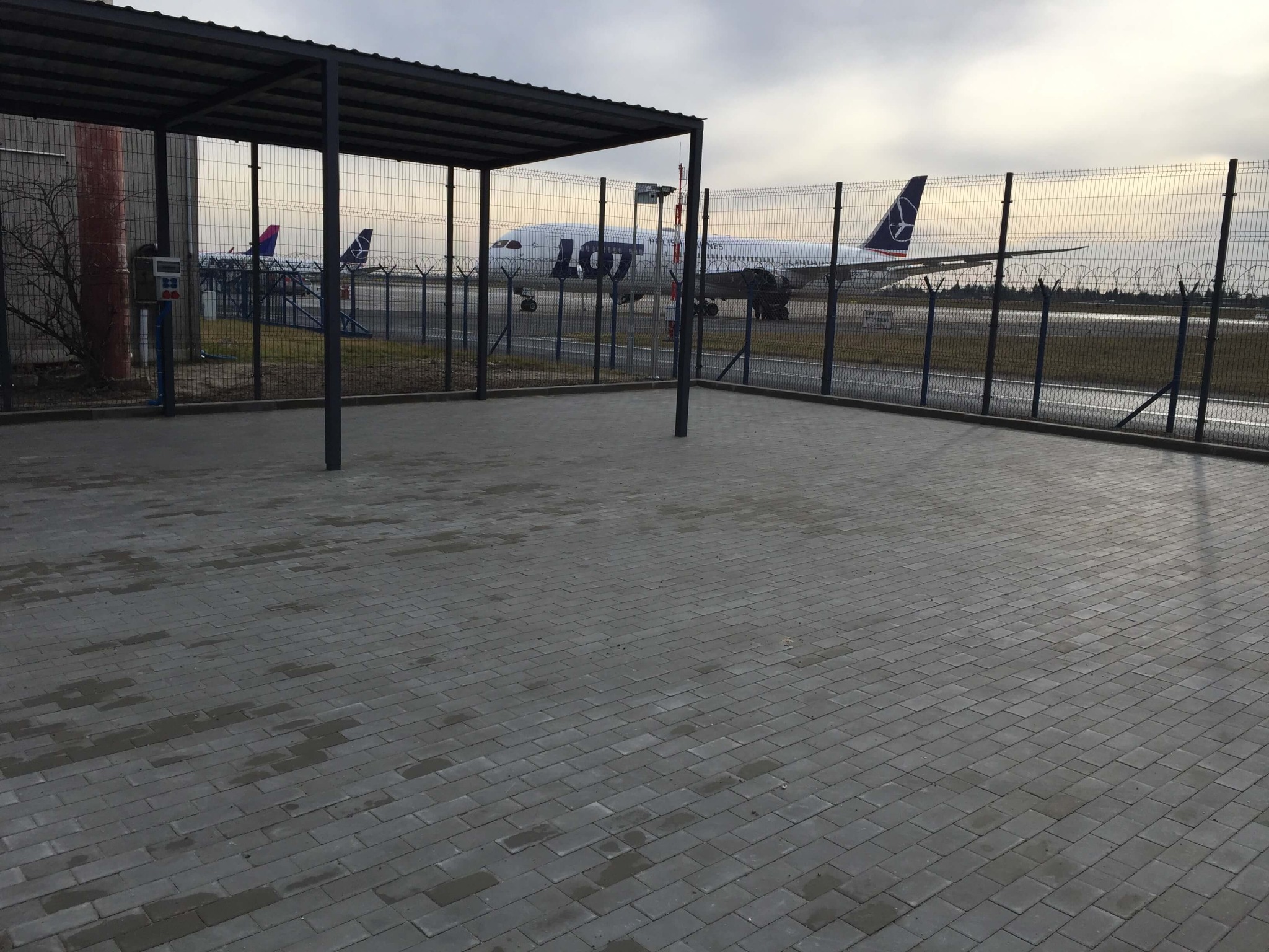 Szara kostka brukowa na lotnisku, w tle samolot LOT-u za ogrodzeniem z drutu kolczastego.