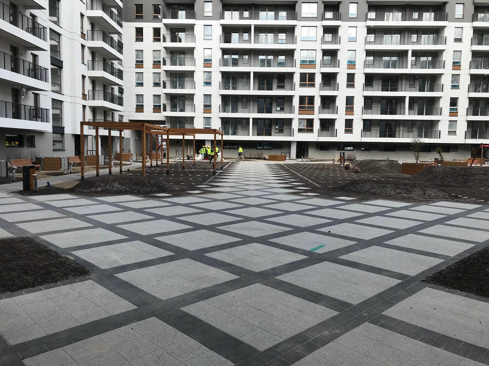 Nowoczesny plac przed budynkiem mieszkalnym z ułożoną szarą kostką brukową w geometryczny wzór, widoczni pracownicy w kamizelkach odblaskowych przygotowujący teren pod nasadzenia.