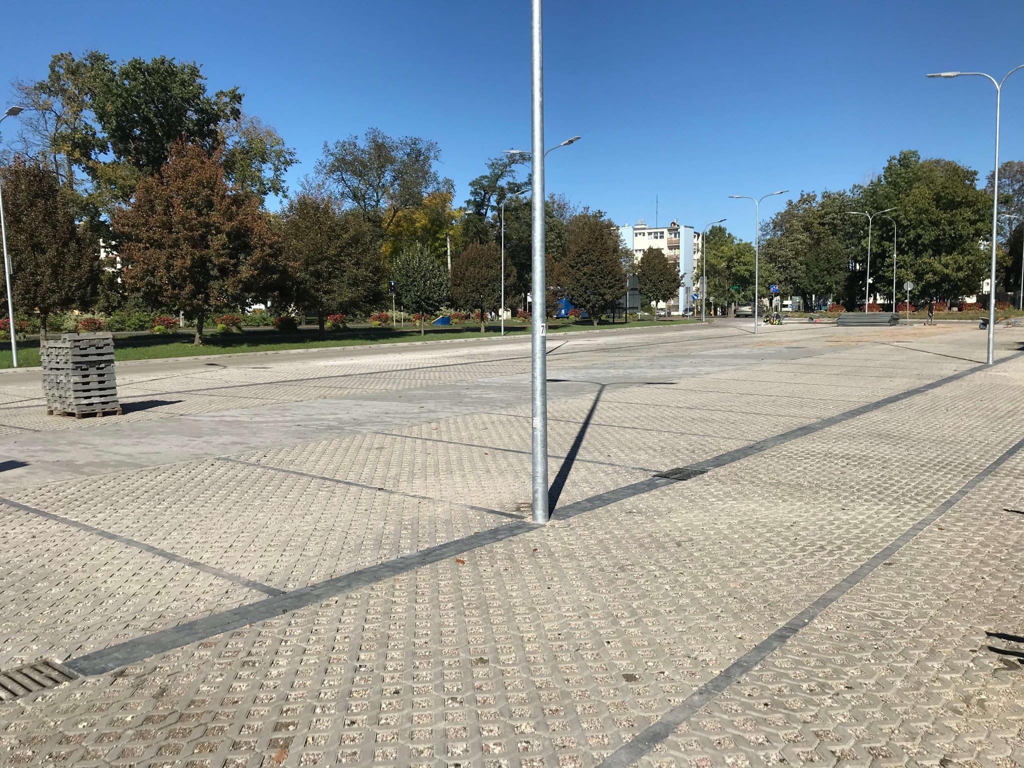 Rozległy plac w Warszawie wyłożony ażurową kostką betonową, z widocznymi liniami drenażowymi i słupami oświetleniowymi, w tle drzewa i budynki.