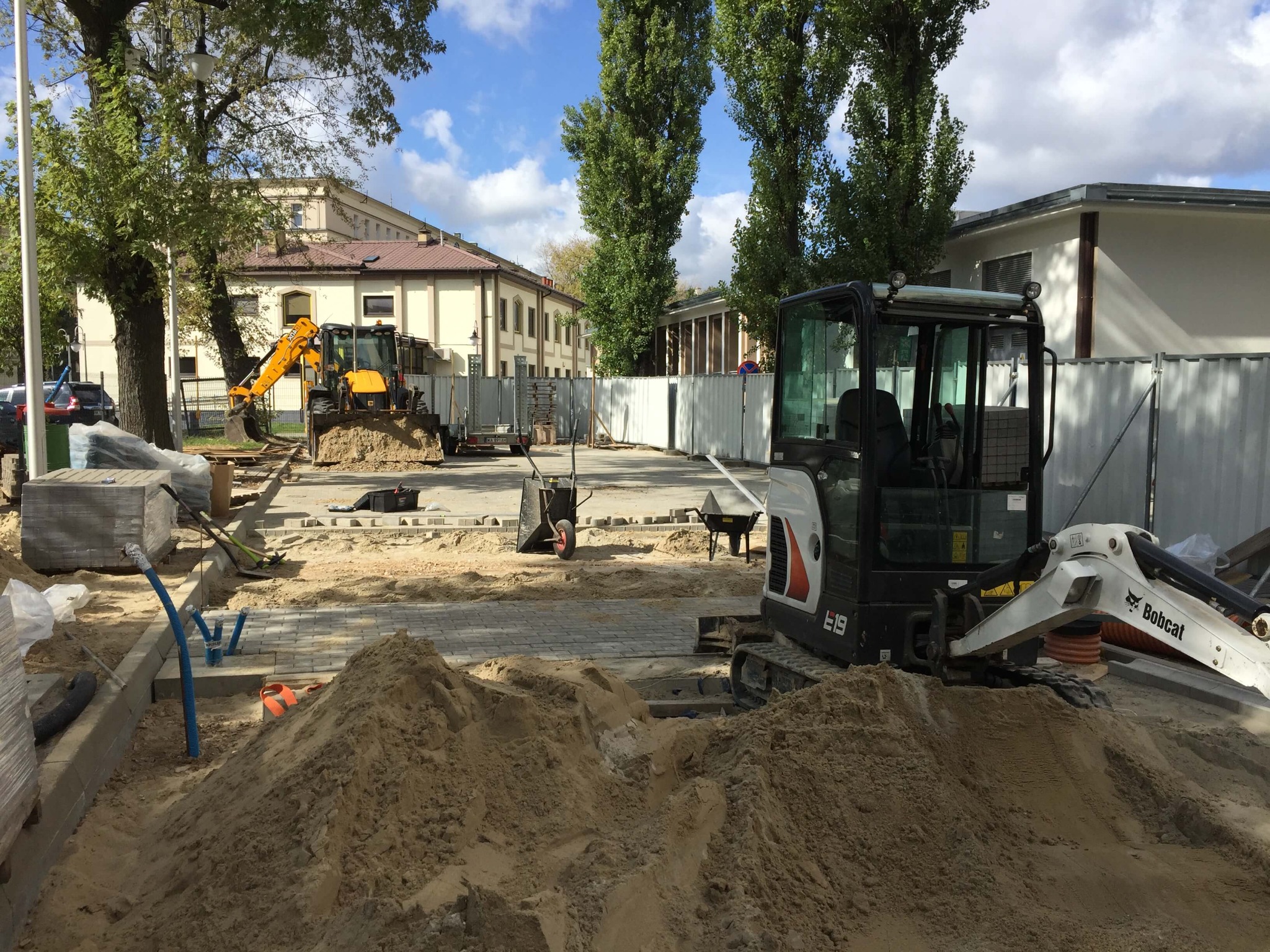 Plac budowy z dwoma minikoparkami (Bobcat i żółta) na tle budynku w trakcie układania szarej kostki, widoczne narzędzia i hałdy piachu.