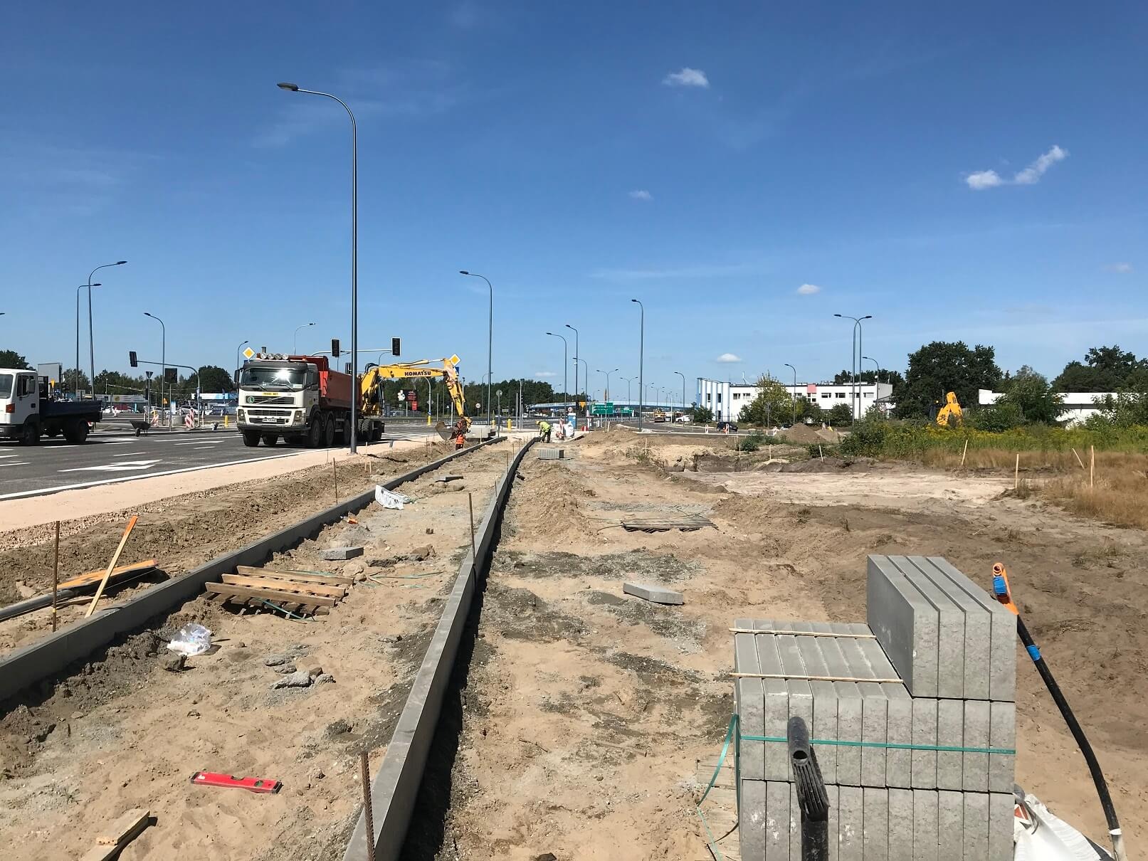 Budowa drogi z kostki brukowej: widok na krawężnik, stos kostki brukowej, ciężarówkę i koparkę Komatsu w tle, na tle błękitnego nieba.