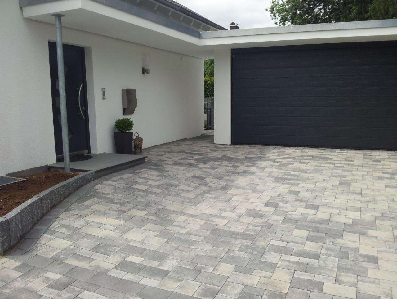 Nowoczesny podjazd z szaro-białej kostki brukowej, prowadzący do domu z garażem i ciemnymi drzwiami wejściowymi. Rabata z ziemią po lewej stronie.