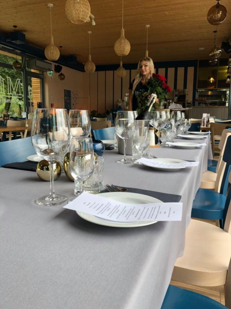 Uroczysty stół z szarą pościelą, nakryty białymi talerzami, kryształowymi kieliszkami i srebrnymi sztućcami, z menu na talerzu, dekoracjami w odcieniach złota i niebieskimi akcentami, w tle...