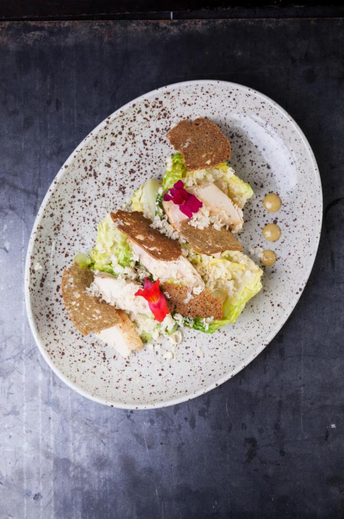 Elegancko podany Cezar z kurczakiem, grzankami z ciemnego pieczywa i jadalnymi kwiatami na talerzu w stylu rustykalnym z ciemnym tłem.