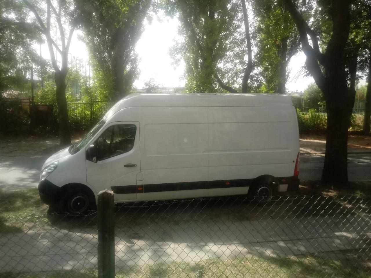 Biały bus dostawczy zaparkowany na ulicy, z widocznym fragmentem ogrodzenia i drzewami w tle, w słoneczny dzień. Widok z boku.