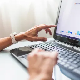 Ręce osoby pracującej na laptopie, wskazującej na wykres słupkowy Agriculture i Manufacturing, biały zegarek na nadgarstku, jasne tło.