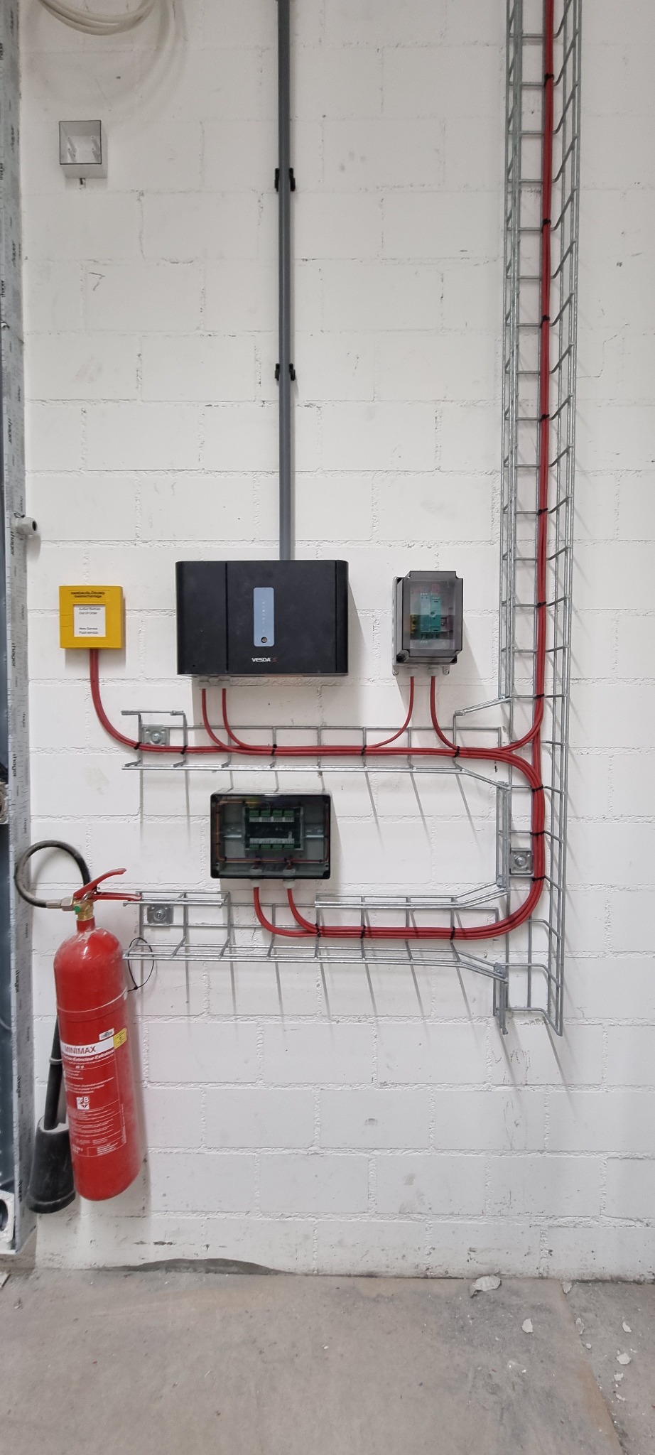 Rozdzielnice elektryczne, przewody w korytach kablowych i gaśnica zamontowane na białej ścianie z bloczków betonowych w pomieszczeniu przemysłowym.