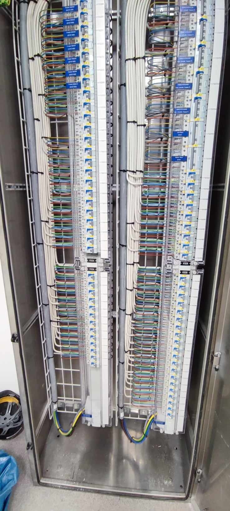 Rozdzielnica elektryczna z uporządkowanymi kablami w różnych kolorach, zakończonymi na listwach zaciskowych z niebieskimi oznaczeniami, umieszczona w metalowej szafie.