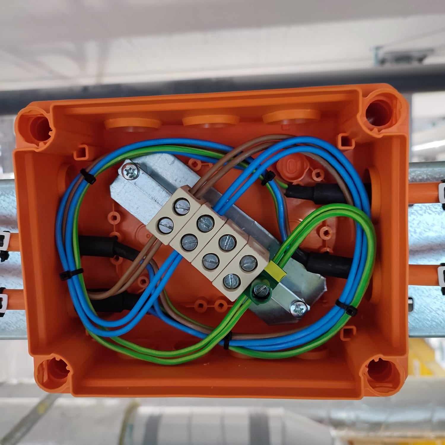 Pomarańczowa puszka instalacyjna z podłączonymi przewodami: niebieskimi, zielonymi i brązowymi, zabezpieczonymi czarnymi opaskami zaciskowymi i złączką listwową z widocznymi śrubami.