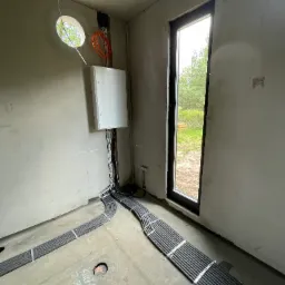 Surowy stan wnętrza z widocznymi, ułożonymi w korytkach kablami elektrycznymi, prowadzącymi do białej skrzynki na ścianie; widoczne okrągłe okno z widokiem na zieleń i duże okno z widokiem...