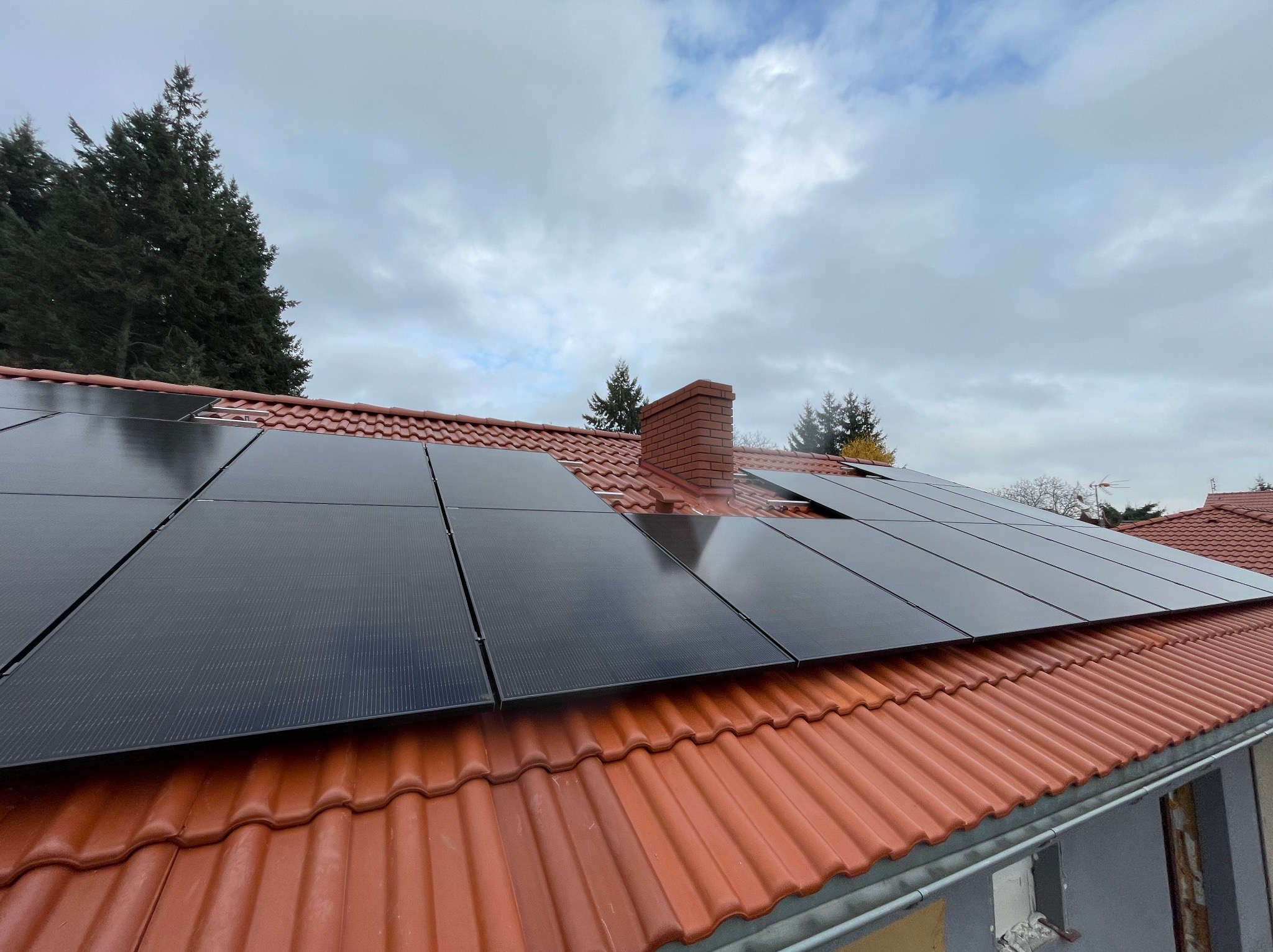 Panele fotowoltaiczne zamontowane na dachu z czerwonej dachówki, widoczny komin z cegły i fragment rynny, w tle drzewa i pochmurne niebo.