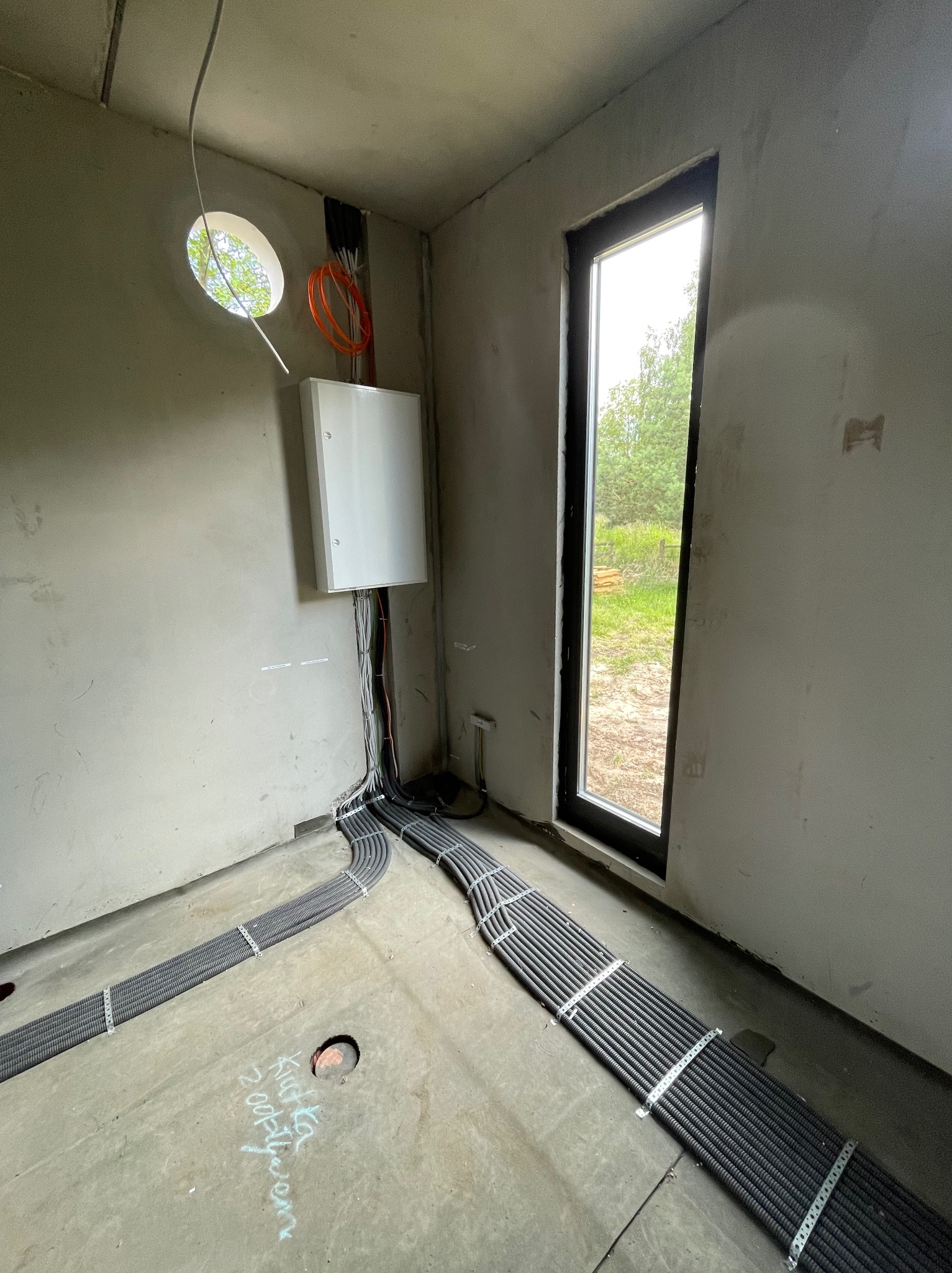 Surowy stan wnętrza z widocznymi, ułożonymi w korytkach kablami elektrycznymi, prowadzącymi do białej skrzynki na ścianie; widoczne okrągłe okno z widokiem na zieleń i duże okno z widokiem...