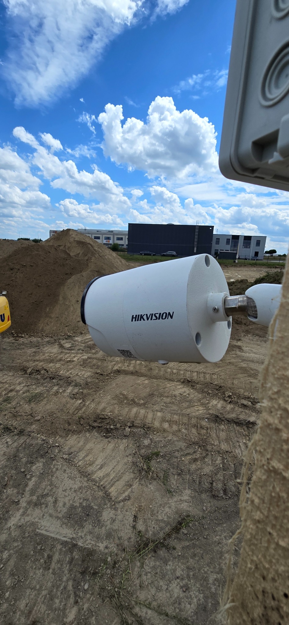 Kamera monitoringu marki Hikvision zamontowana na zewnątrz, widok na plac budowy z hałdami ziemi i budynkami w tle, błękitne niebo z chmurami.