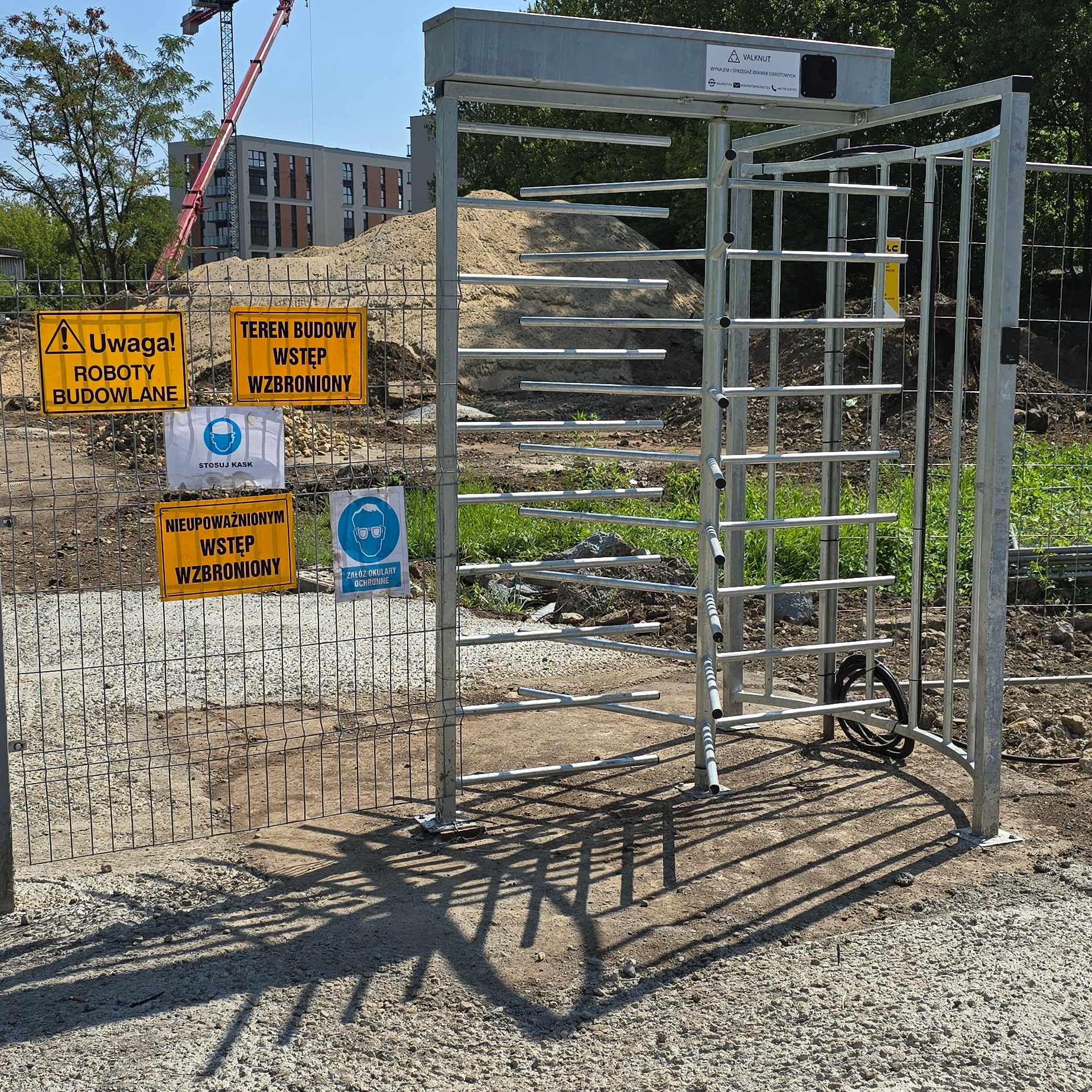 Metalowa bramka obrotowa na terenie budowy, z widocznymi tablicami informacyjnymi o zakazie wstępu i konieczności stosowania środków ochrony osobistej, w tle budynek w trakcie budowy i żuraw.