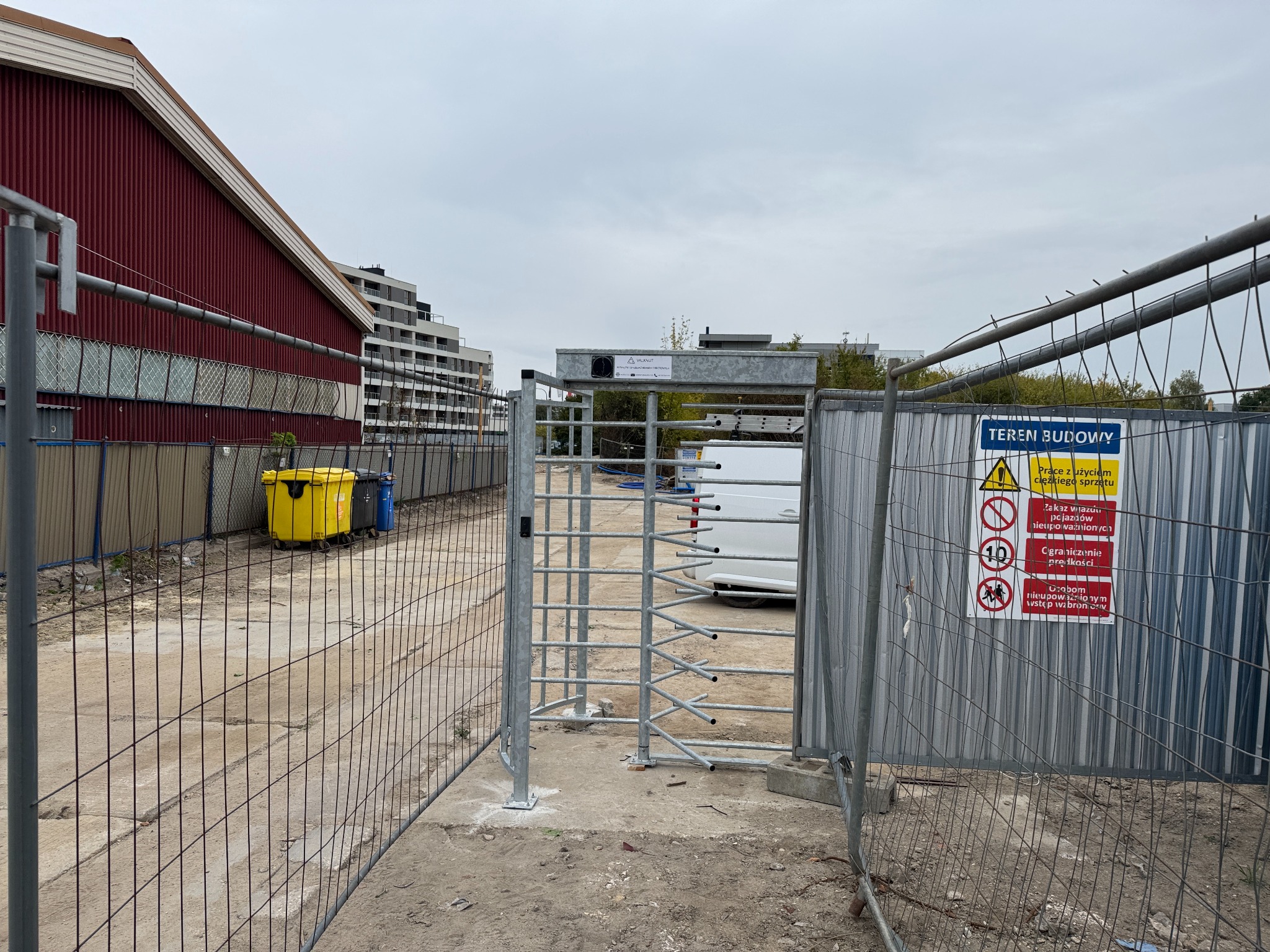 Teren budowy z metalowym ogrodzeniem i bramką obrotową, widoczne znaki ostrzegawcze, w tle budynki i żółte kontenery na śmieci.