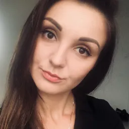 Portret kobiety z długimi brązowymi włosami, ubranej w czarną marynarkę, z delikatnym makijażem i złotym łańcuszkiem z zawieszkami