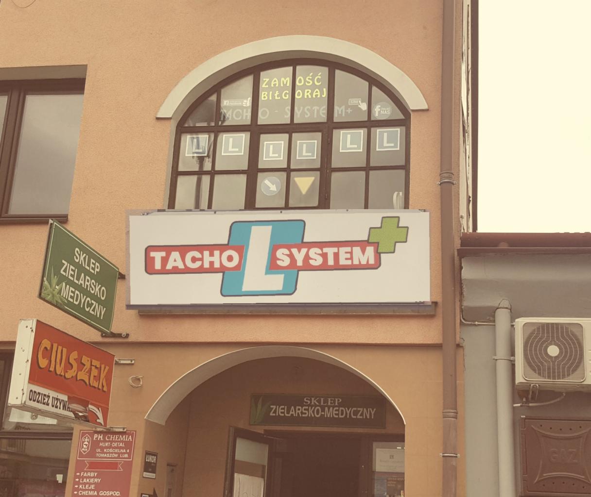 Fasada budynku z szyldem firmy Tacho System z literą L w kwadracie, okno z naklejonymi literami L i strzałką w dół, oraz szyldy innych sklepów: zielarsko-medycznego i odzieży używanej.