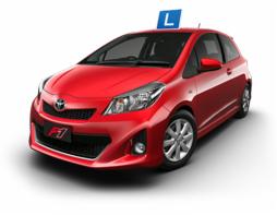 Czerwony samochód Toyota Yaris z niebieską literą 'L' na dachu, widok z przodu, lekko z góry, na białym tle.