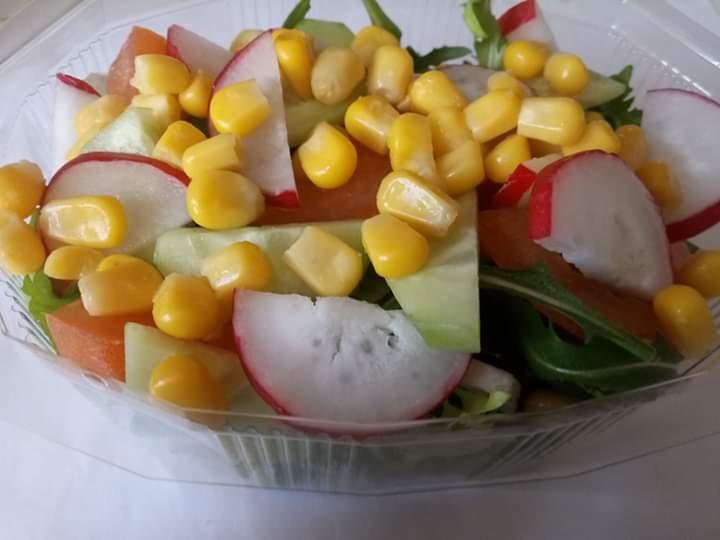 Świeża sałatka warzywna z kukurydzą, rzodkiewką, ogórkiem i marchewką w przezroczystym plastikowym pojemniku, widoczne liście sałaty, ujęcie z góry.