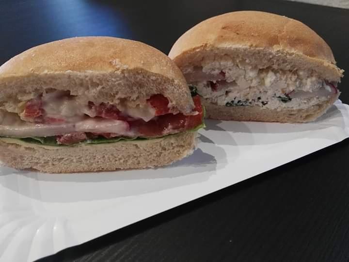 Dwa okrągłe, domowe burgery na białej papierowej tacy: jeden z pomidorami, serem i sałatą, drugi z pastą twarogową z ziołami.