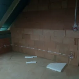 Wnętrze poddasza w trakcie budowy z widoczną instalacją elektryczną w ścianie z cegieł dziurawki oraz izolacją stropu i fragmentami styropianu na podłodze.