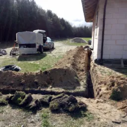 Wąski wykop w ziemi obok budynku w trakcie budowy, z widocznymi kablami wewnątrz, przyczepą i stertą ziemi w tle.