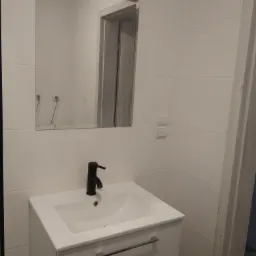 Nowoczesna łazienka z białą umywalką nablatową, czarną baterią i dużym lustrem z oświetleniem punktowym, białe płytki na ścianach.