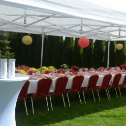 Ustawiony stół pod białym namiotem plenerowym, z czerwonymi krzesłami, białym obrusem, zastawą, dekoracjami w postaci czerwonych i żółtych lampionów oraz kwiatów, z dystrybutorem do napojów...
