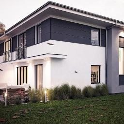 Castor Pracownia Projektowa Agata Sobkowiak - Dwukondygnacyjny dom z elementami grafitowej elewacji, balkonem z czarną balustradą i oświetlonym tarasem z drewnianymi meblami ogrodowymi, otoczony dekoracyjną trawą i jesiennymi liśćmi na trawniku.