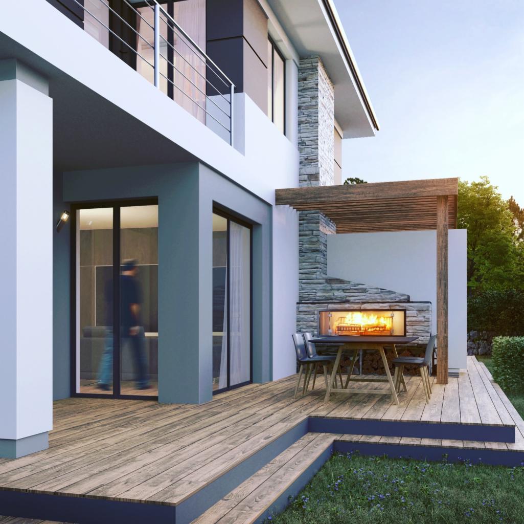 Nowoczesny dom z tarasem z drewna, na którym znajduje się kamienny kominek z paleniskiem, stół i krzesła. W tle widoczny ogród i osoba przechodząca przez przeszklone drzwi.