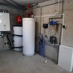 Kotłownia z dwoma białymi zbiornikami akumulacyjnymi, czerwonym naczyniem wzbiorczym i wiszącym kotłem gazowym Panasonic na surowej betonowej ścianie.