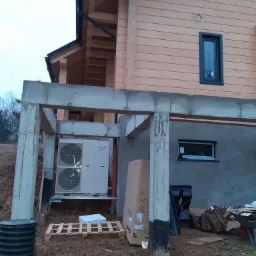 Zewnętrzna jednostka dwuwentylatorowej pompy ciepła Panasonic Aquarea zamontowana na zewnątrz budynku w trakcie instalacji, widoczne elementy konstrukcyjne budynku, paleta i karton po urządzeniu.