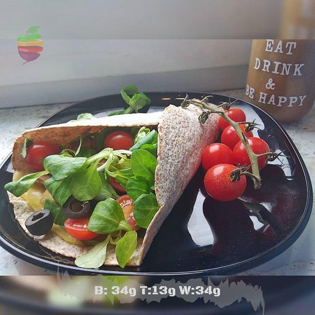 Tortilla z sałatką, pomidorami koktajlowymi i oliwkami na czarnym talerzu obok kubka z napisem 'Eat Drink & Be Happy', widoczne wartości odżywcze B:34g T:13g W:34g.