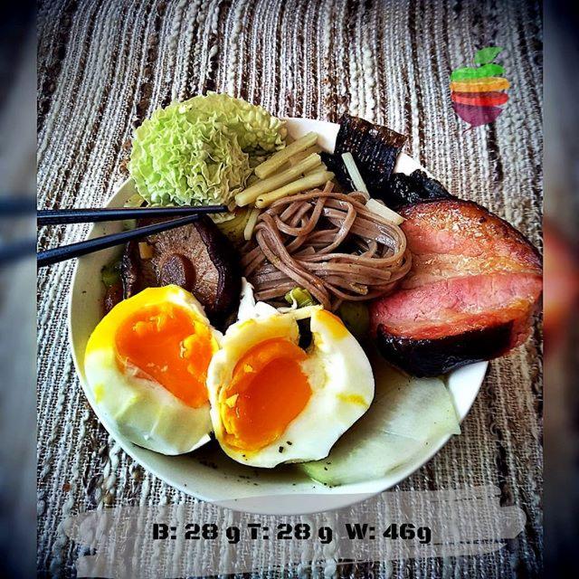Kompozycja na talerzu: przekrojone jajko na miękko, soba, wodorosty nori, mięso, warzywa i pałeczki na tle teksturowanej serwety. Makroelementy wypisane na dole zdjęcia.