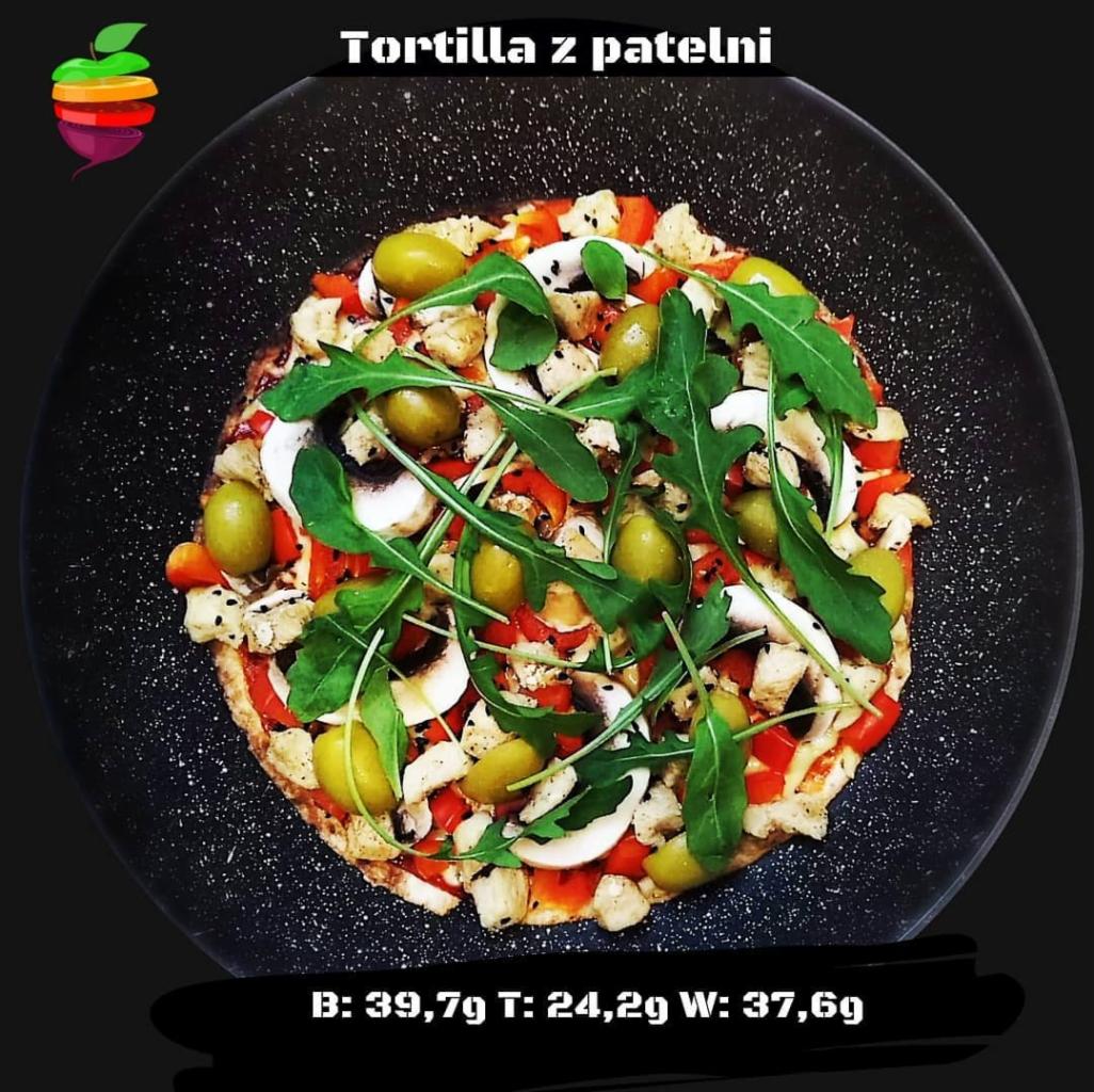 Tortilla z patelni na czarnym talerzu, udekorowana rukolą, oliwkami, pieczarkami i papryką, z informacją o wartościach odżywczych na dole.