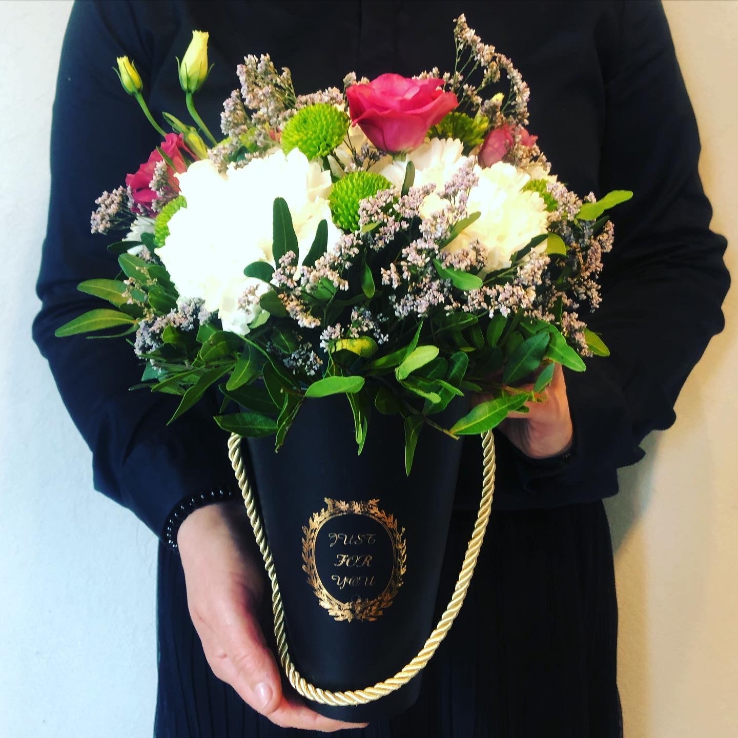 Kompozycja kwiatowa w czarnym pudełku z napisem 'Just for you' trzymana przez osobę w czarnej bluzce, z widocznymi różowymi i białymi różami, zielonymi chryzantemami i drobnymi fioletowymi kwiatami.