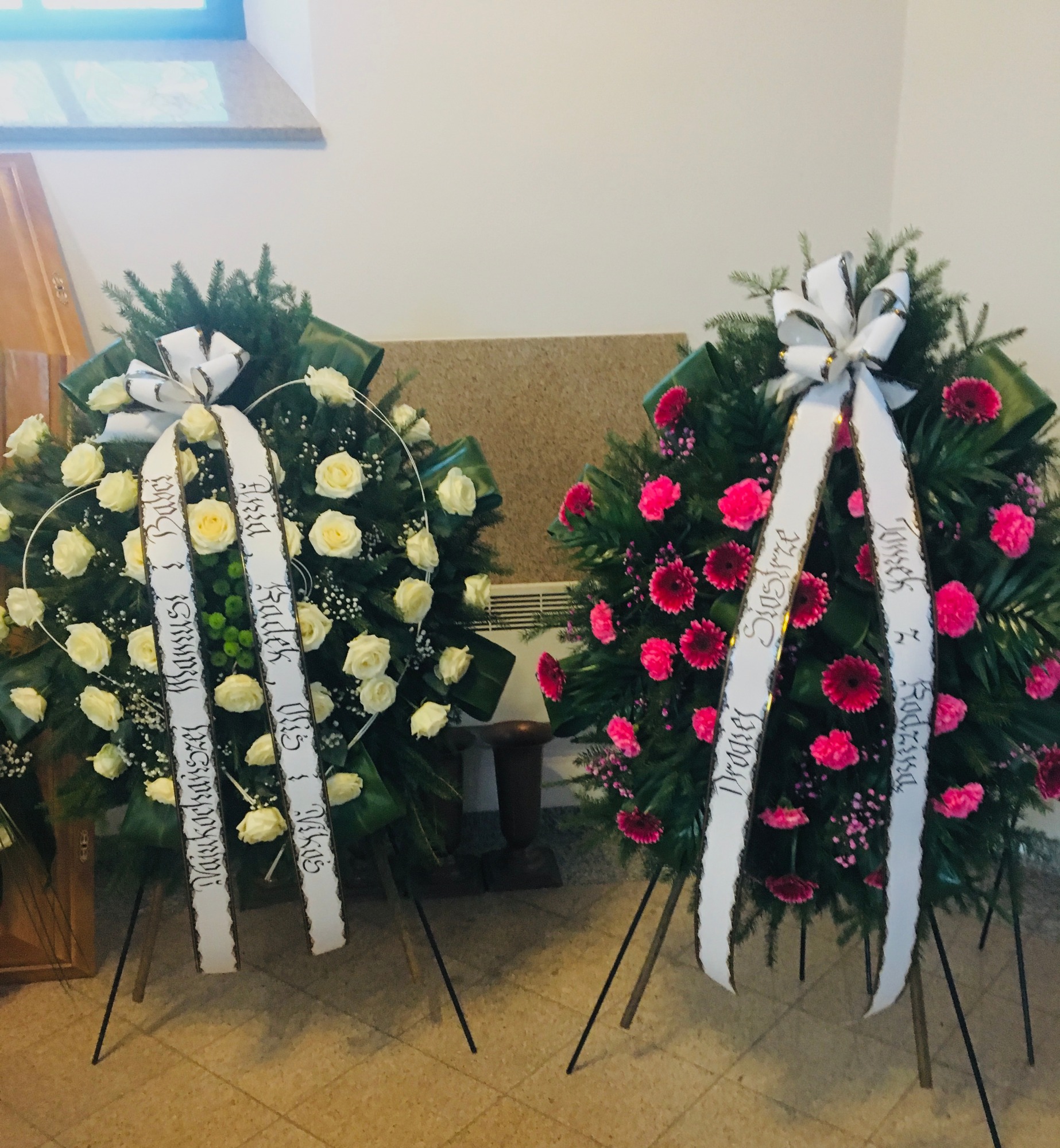 Dwa wieńce pogrzebowe na stojakach: jeden z białych róż, drugi z różowych kwiatów, każdy z białą szarfą z czarnym napisem, ustawione w jasnym wnętrzu.