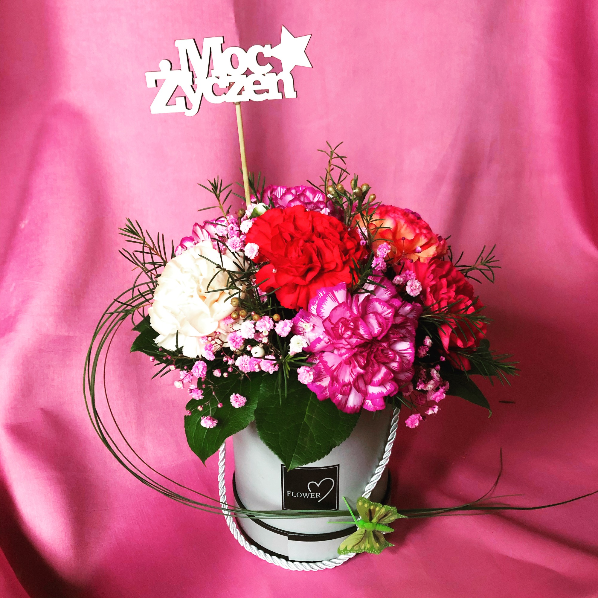 Kompozycja kwiatowa w okrągłym pudełku z napisem 'Moc Życzeń' i dekoracyjną motylkową ozdobą, na różowym tle.