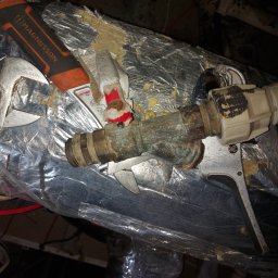 HCM-AirControl - Zardzewiałe, mosiężne rury połączone z białą plastikową złączką, naprawiane za pomocą klucza nastawnego i taśmy izolacyjnej, na tle izolacji z folii aluminiowej.