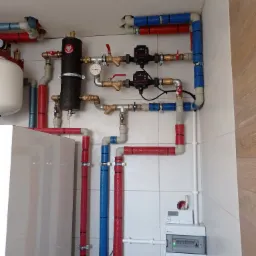 Instalacja grzewcza z rurami w kolorze czerwonym i niebieskim, zaworami, pompami obiegowymi i zbiornikiem wyrównawczym w kotłowni w Skierniewicach, widoczny również panel sterowania.