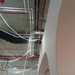 HCM-AirControl - Widok na sufit w trakcie montażu instalacji: rury w izolacji aluminiowej, czerwone rury instalacji przeciwpożarowej, czarne rury wentylacyjne, białe przewody elektryczne, drabiny monterskie, łukowe...