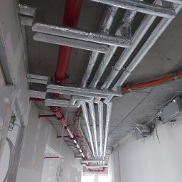HCM-AirControl - Widok instalacji wentylacyjnej i przeciwpożarowej pod sufitem w budynku w trakcie wykończenia, widoczne rury izolowane folią aluminiową, czerwone rury przeciwpożarowe, kable i pracownik w kasku.