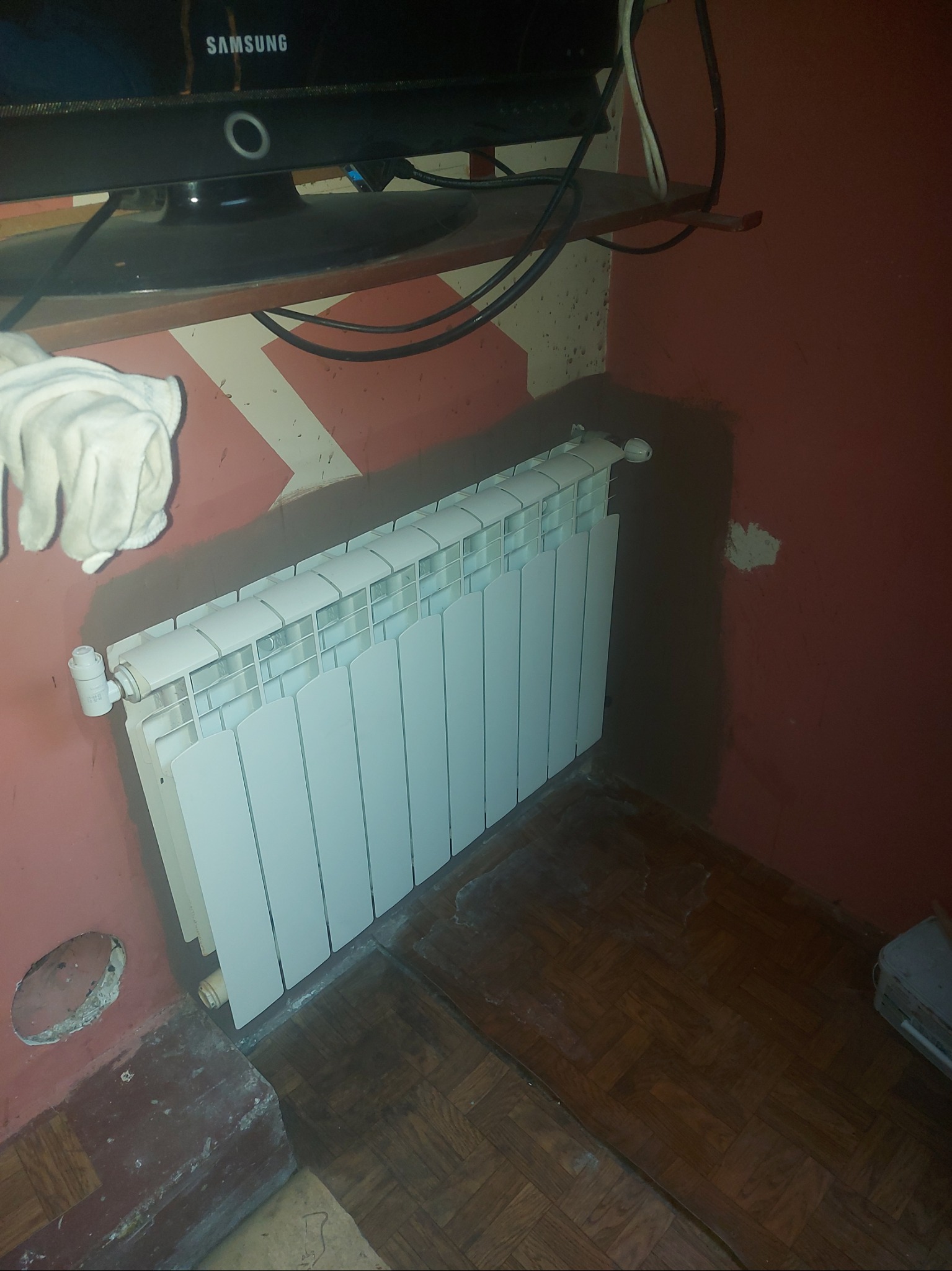 Biały grzejnik panelowy zamontowany na czerwonej ścianie z widocznymi śladami po remoncie, nad nim półka z telewizorem Samsung, na podłodze parkiet.