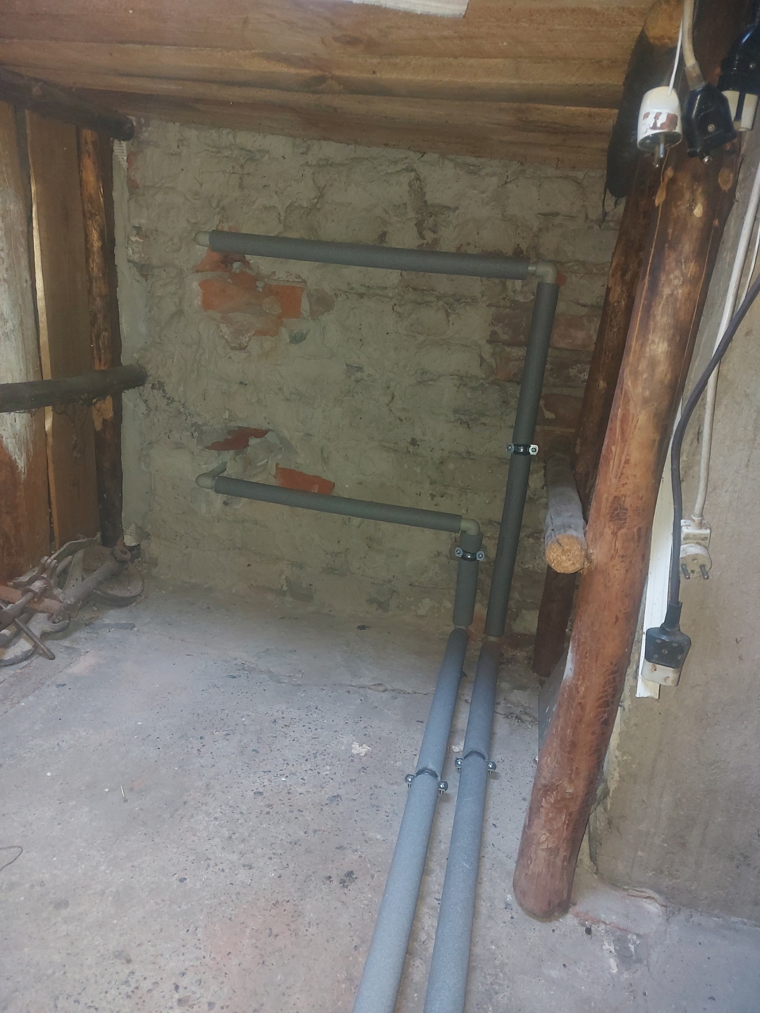 Szare rury instalacji grzewczej z izolacją, zamocowane do ściany w surowym pomieszczeniu z drewnianym stropem i widocznymi cegłami.