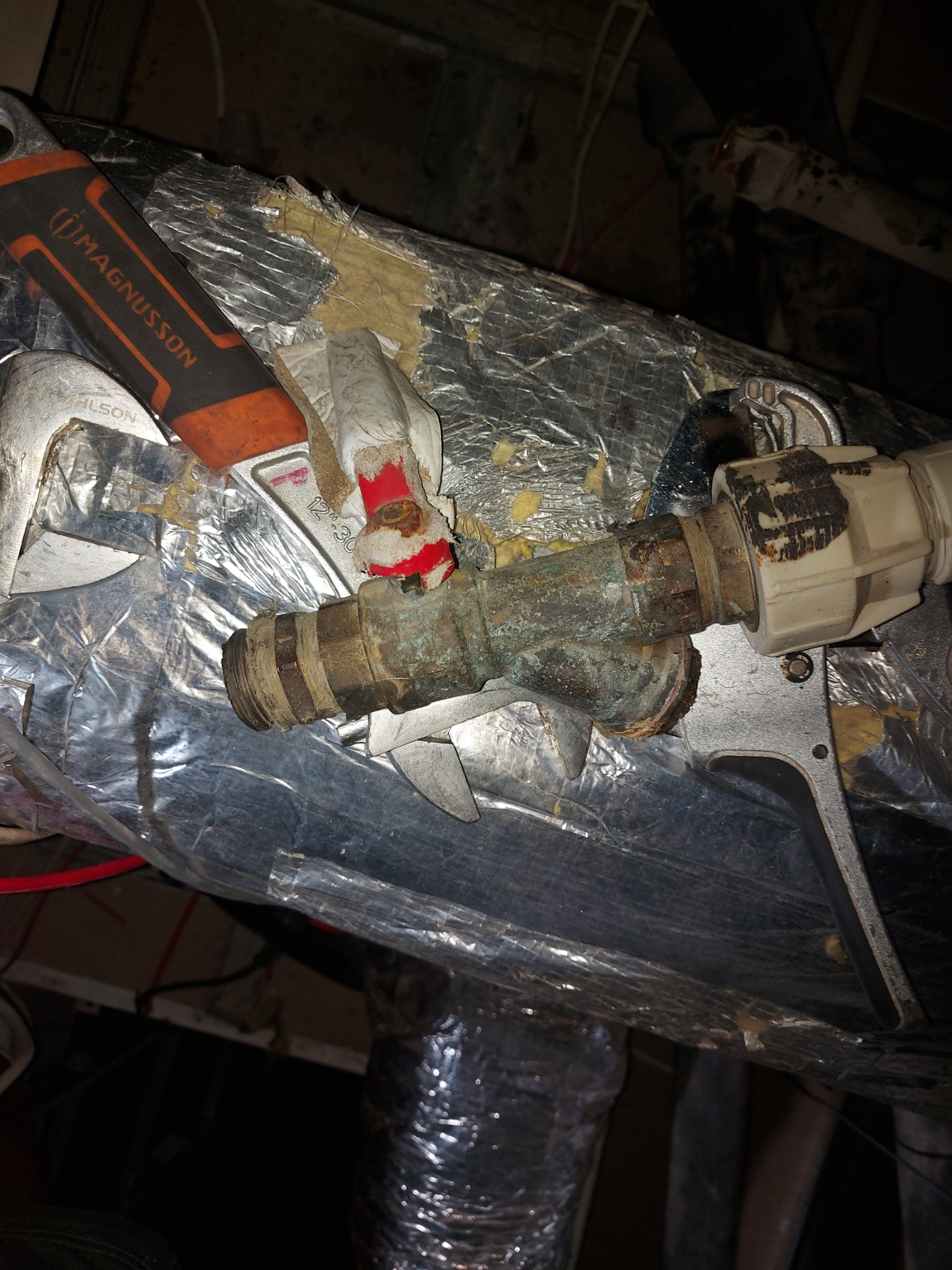 Zardzewiałe, mosiężne rury połączone z białą plastikową złączką, naprawiane za pomocą klucza nastawnego i taśmy izolacyjnej, na tle izolacji z folii aluminiowej.